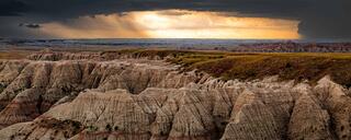 Fine art panoramic landscape photograph of the Badlands with layered rock formations beneath a dramatic golden storm sky.