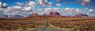 Long straight road leading into vast Utah desert, framed by red rock mesas and dramatic clouds under open sky.