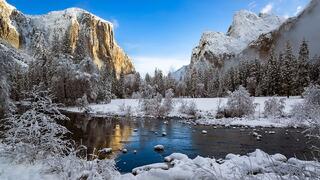 A serene winter landscape showcases snow-covered trees, icy waters, and towering granite cliffs bathed in soft light.