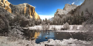 A serene winter landscape showcases snow-covered trees, icy waters, and towering granite cliffs bathed in soft light.