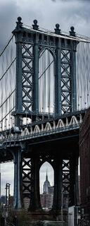 View beneath the Manhattan Bridge in DUMBO, with towering steel arches, suspension cables, and the Empire State Building framed below.
