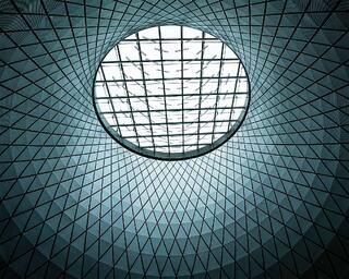 Looking up through a geometric glass dome, repeating triangular patterns curve toward a circular opening filled with soft, even light.