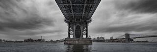 A black-and-white photograph looking up beneath the Manhattan Bridge, where massive steel supports frame the river below.