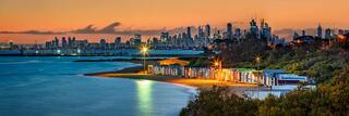 The Brighton Bathing Boxes lit at dusk on Dendy Street Beach with the Melbourne skyline across Port Phillip Bay, Victoria, Australia.