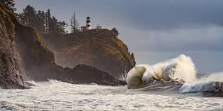 A large wave breaking below the Cape Disappointment Lighthouse on its forested headland during a king tide, Washington.