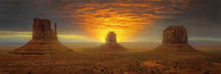 West Mitten, East Mitten, and Merrick Butte in Monument Valley at sunrise, the sun rising between the buttes beneath an orange, cloud-streaked sky.