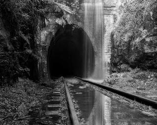 A waterfall pouring over the brick portal of a disused railway tunnel with flooded tracks leading into the dark, black and white, Helensburgh, New South Wales