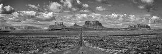 Long straight road leading into vast Utah desert, framed by red rock mesas and dramatic clouds under open sky.