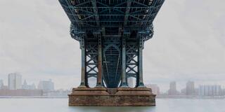 Symmetrical view beneath a New York City bridge with soft light, muted blue tones, still water, and a calm, misty atmosphere.