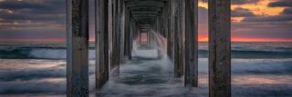 View beneath a coastal pier at sunset in San Diego, with repeating concrete columns framing flowing ocean waves and warm light glowing along the horizon