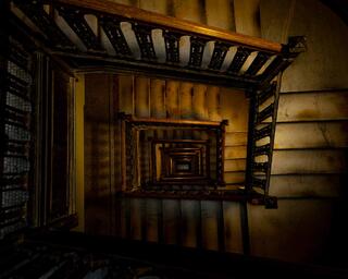 Looking down a historic stairwell, repeating railings and steps form a geometric spiral lit by warm light, evoking quiet reflection.
