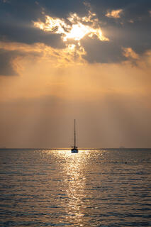 A single sailboat anchored in silhouette on calm water at sunset off Railay Beach, Thailand, with sunlight breaking through clouds above.