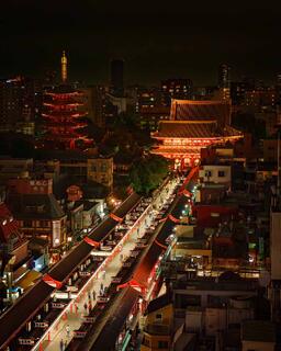Elevated night view of Nakamise-dori leading to the lit Hozomon gate and Senso-ji in Asakusa, Tokyo, with a 5-story pagoda at left and the city skyline behind.