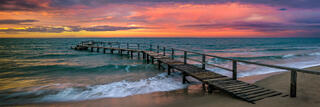 A weathered timber jetty running from the sand into the sea beneath a pink and orange sunset sky, Shelley Beach, Portsea, Victoria.
