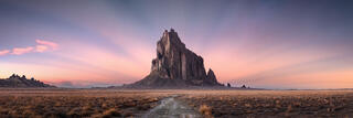 Shiprock, an isolated volcanic rock formation on a flat desert plain in northwestern New Mexico, with a dirt track leading toward it under a pink & purple dusk.