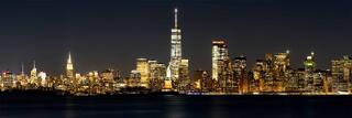 Night view of the New York City skyline with One World Trade Center illuminated, the Statue of Liberty centered, and dark water in the foreground.