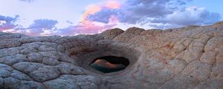 Natural sandstone basin holding still water that reflects the sky, surrounded by sculpted rock in a quiet desert landscape.