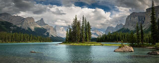 Spirit Island in Jasper National Park surrounded by turquoise lake water, evergreen forest, Canadian Rockies, and dramatic mountain clouds.