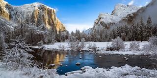 A serene winter landscape showcases snow-covered trees, icy waters, and towering granite cliffs bathed in soft light.