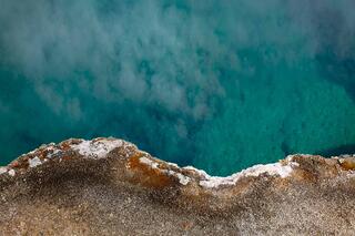 The edge of a Yellowstone hot spring where turquoise water meets rust and white mineral crust, with steam across the surface.