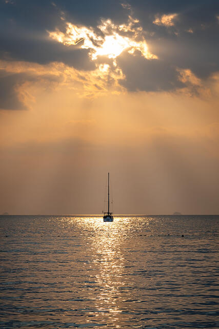 A lone sailboat sits anchored on calm golden water off Railay Beach, Thailand, at sunset, its mast rising vertically into a dramatic sky where crepuscular rays