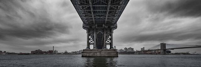 A black-and-white photograph looking up beneath the Manhattan Bridge, where massive steel supports frame the river below.