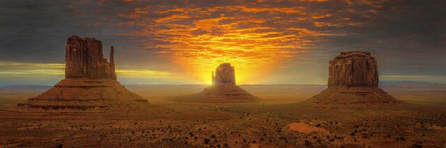 The image depicts three prominent rock spires in the vast desert, set against a dramatic sky filled with hues of orange, pink, and purple as the sun rises.