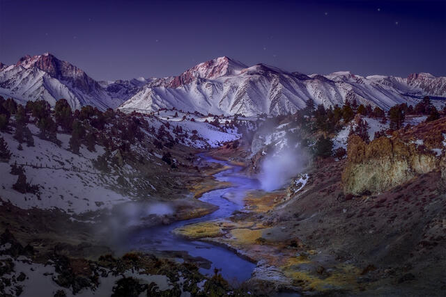 A winter landscape with a gently flowing river and rising steam, framed by snow covered mountains and a deep blue sky, creating a quiet and contemplative scene.
