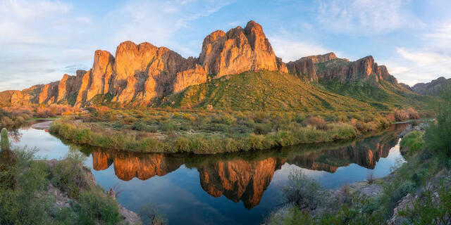 Fine art panoramic landscape photograph of the Goldfield Mountains reflected in the still waters of the Salt River at sunrise, with lush riparian vegetation.