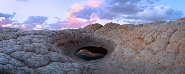 Natural sandstone basin holding still water that reflects the sky, surrounded by sculpted rock in a quiet desert landscape.