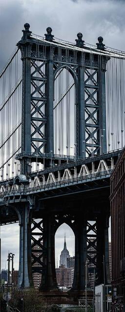 View beneath the Manhattan Bridge in DUMBO, with towering steel arches, suspension cables, and the Empire State Building framed below.