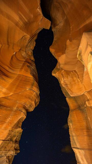 Carved sandstone walls of Antelope Canyon seen from below, with a star-filled night sky visible through the narrow opening above, Page, Arizona.