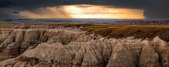 Eroded Badlands buttes and mixed-grass prairie lit by a break in a storm at sunset, Badlands National Park, South Dakota.