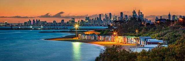 The Brighton Bathing Boxes lit at dusk on Dendy Street Beach with the Melbourne skyline across Port Phillip Bay, Victoria, Australia.