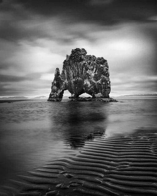 The basalt sea stack Hvitserkur standing offshore at low tide with rippled sand in the foreground, black and white, Vatnsnes peninsula, Iceland.