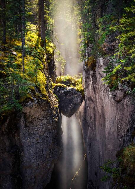 A waterfall falling beneath a heart-shaped chockstone wedged in the narrow slot of Maligne Canyon, with mist and morning light between mossy walls, Jasper NP