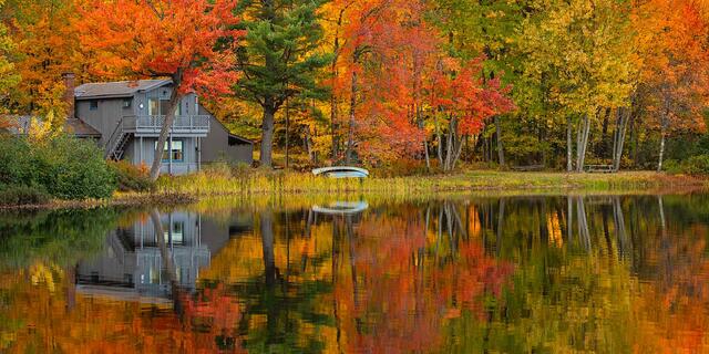 A lakeside house among autumn hardwoods with an overturned canoe on the shore, mirrored in still water, eastern New Hampshire.