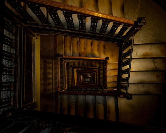 Looking down a historic stairwell, repeating railings and steps form a geometric spiral lit by warm light, evoking quiet reflection.
