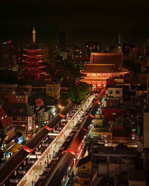 Elevated night view of Nakamise-dori leading to the lit Hozomon gate and Senso-ji in Asakusa, Tokyo, with a 5-story pagoda at left and the city skyline behind.