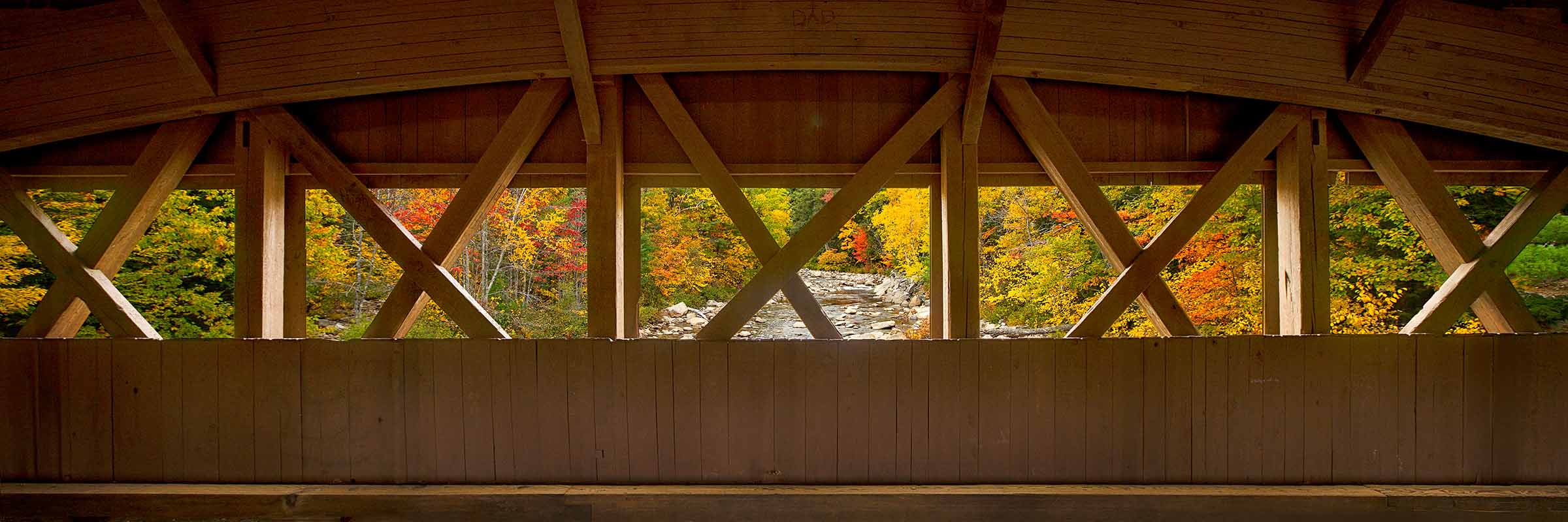 Sheltered View ARTIST’S STORY There is a particular kind of seeing that only becomes possible when you are held. The covered bridges of New Hampshire's White Mountains were built for practical reasons, to protect the wooden decking from the weather, to extend the life of a structure that would otherwise surrender to the seasonal extremes of a New England winter. But step inside one on an autumn morning when the forest on either side of the river has reached its fullest expression of color, and something unexpected happens. The bridge stops being infrastructure and becomes something closer to a frame. Or perhaps a sanctuary. I stopped in the middle of this bridge and turned to face the side. What the timber framework had done to the view outside was extraordinary. The crossed beams and vertical supports had divided the autumn forest into a series of individual panels, each one containing its own arrangement of red and orange and gold, the rocky stream threading through the center panel like a path leading deeper into the color. The cathedral arc of the roof curved above it all, enclosing the space with a warmth that the season outside, for all its brilliance, could not quite match. There is something deeply restorative about the experience of being sheltered while simultaneously connected to something vast and beautiful. The research on psychological restoration tells us that nature environments replenish our depleted attention and reduce the physiological markers of stress. But what this bridge offered was something more specific than that. It offered the particular comfort of enclosure, of being held within a warm and human made space, while the full extravagance of the natural world remained completely visible and completely present just beyond the timber frame. We spend so much of our lives either fully exposed to everything or fully insulated from it. The covered bridge offers a third option. Sheltered and present simultaneously. Protected enough to truly see. Sheltered View is an invitation to find the covered bridge in your own life. The place where you are held just enough to be fully open. Where the frame around the beauty makes the beauty more beautiful. Where being inside and outside at the same time feels, for once, exactly right. Limited Fine Art Edition Each image is released in limited editions, with edition size set by presentation and scale, so the most substantial works remain the most rare.