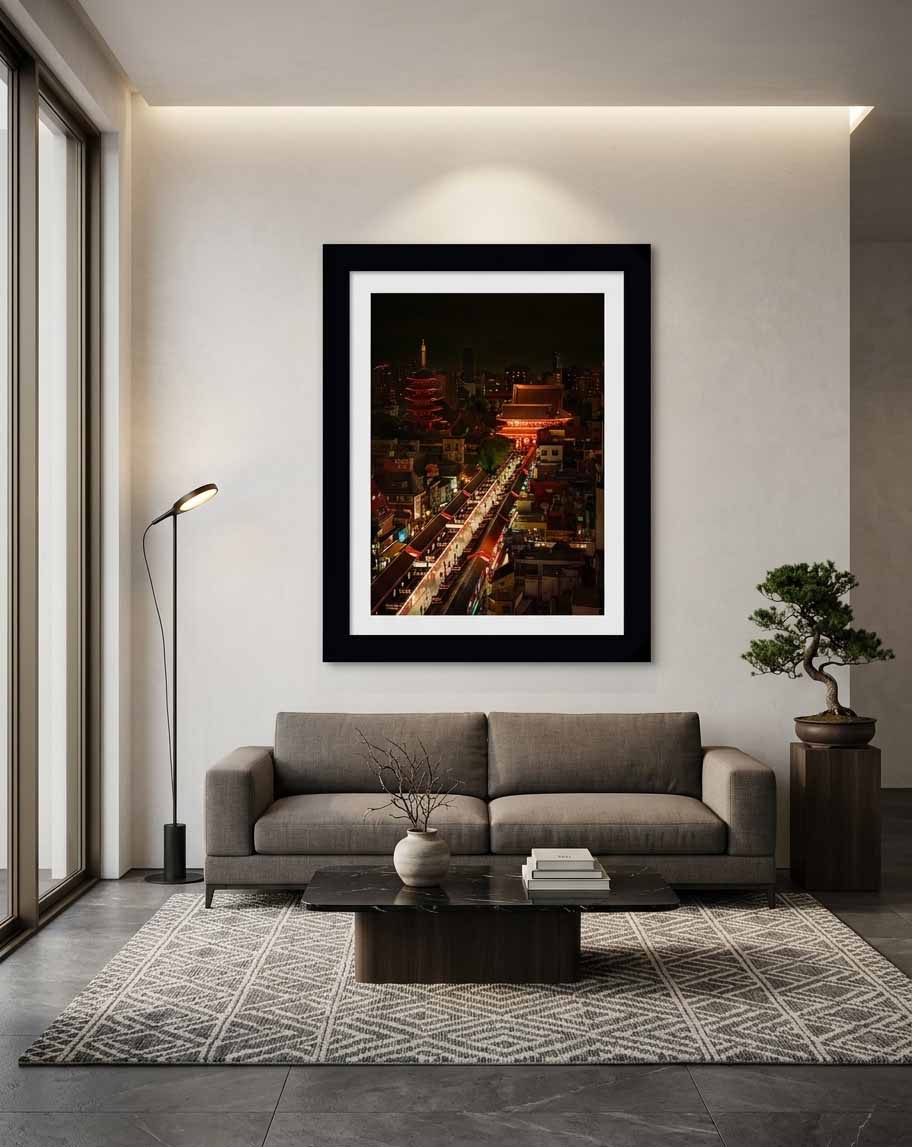 Framed print of Procession, a night view of Senso-ji temple and the lantern-lit Nakamise-dori shopping street in Asakusa, Tokyo, in a black frame with a white mat, hanging on the pale wall of a minimalist modern living room above a gray sofa, with a floor lamp to the left and a bonsai on a wooden pedestal to the right.