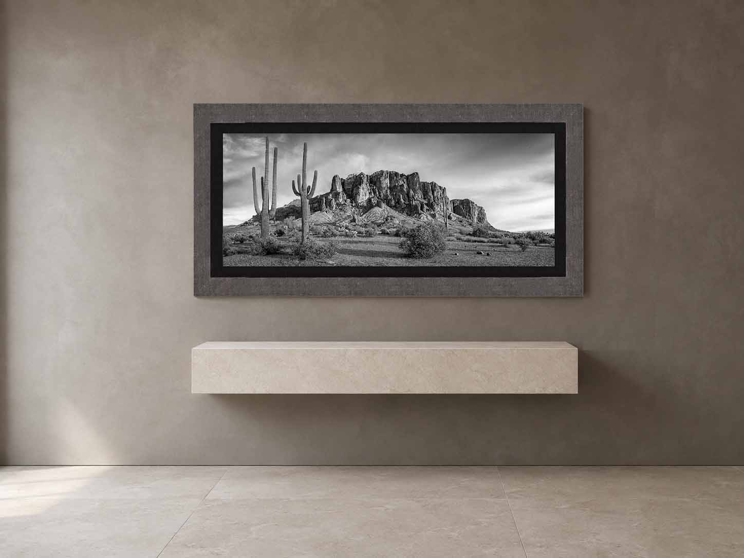 A black and white photograph of saguaros below the Superstition Mountains in Arizona, framed in gray shagreen with a black liner, above a limestone console.