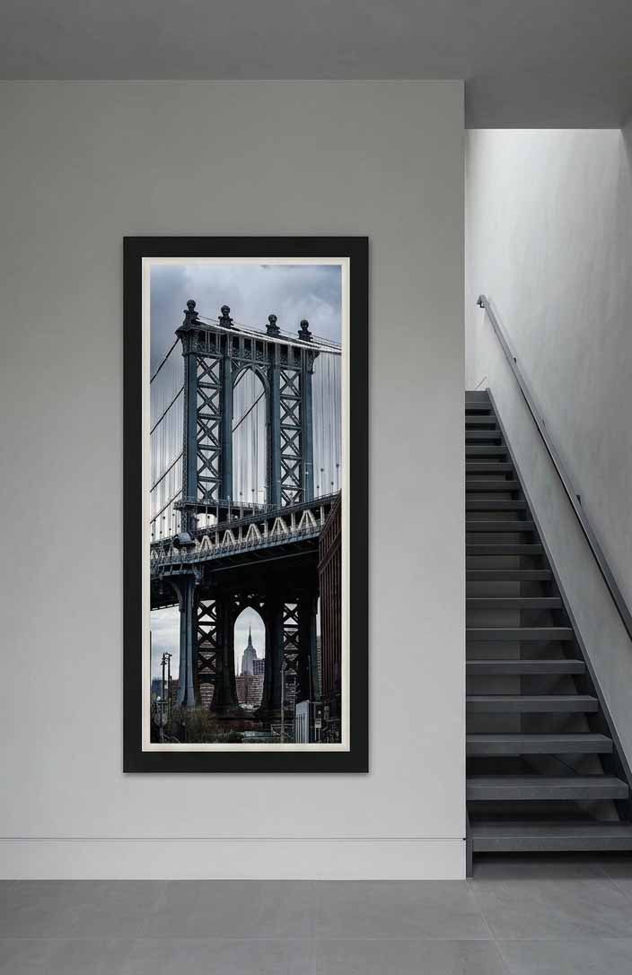 The Manhattan Bridge tower with the Empire State Building in its arch, framed in black with a white liner beside a stair in a gray hall