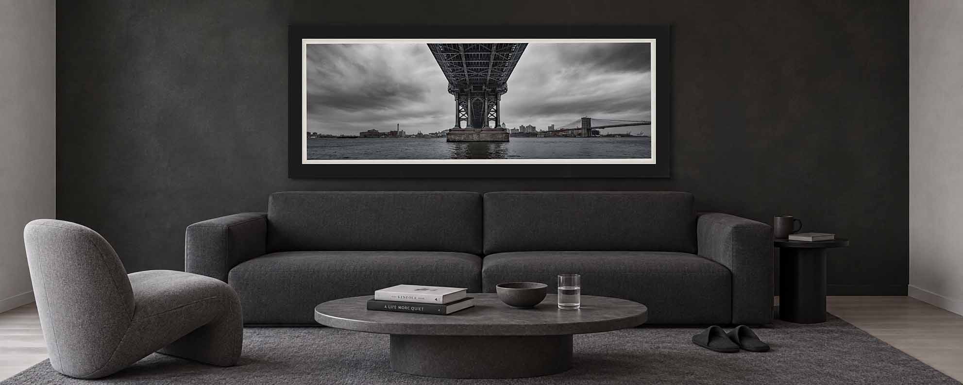 The underside of the Manhattan Bridge over the East River with the Brooklyn Bridge beyond, framed in satin black with a white linen liner above a sofa in a charcoal living room
