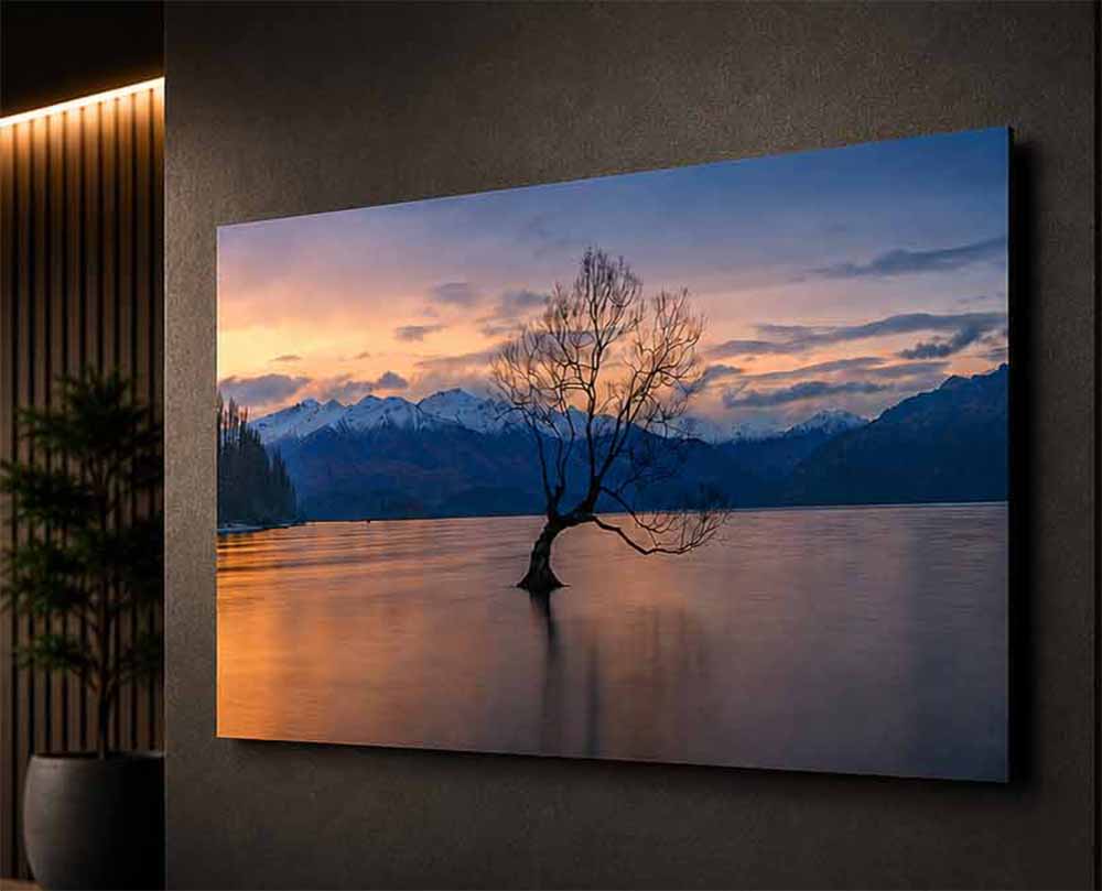 A ChromaLuxe metal print of a lone tree at sunrise on a mountain lake, floating from a dark wall beside a vertical light fixture.