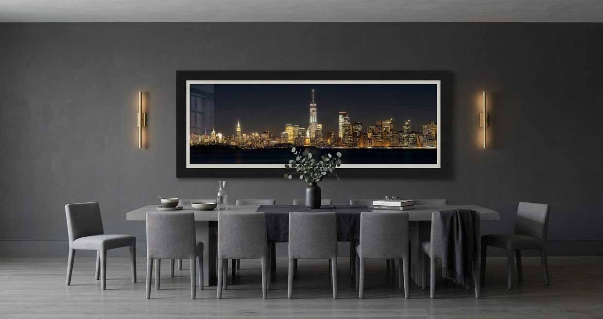 The Lower Manhattan skyline at night across New York Harbor, framed in black with a white linen liner above a stone table in a dark dining room