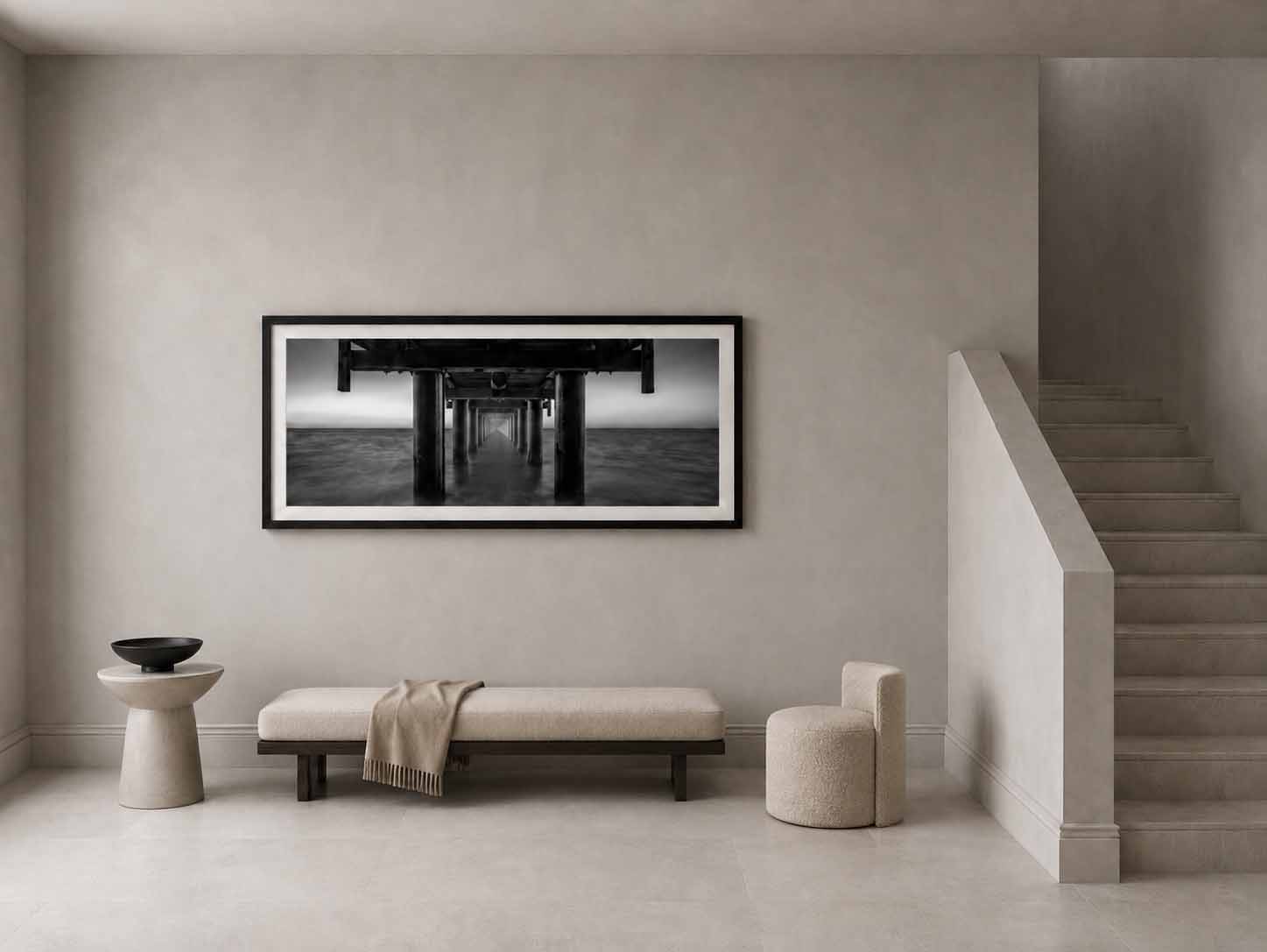 The underside of Urangan Pier in black and white, framed in black with a white mat above a pale bench in a quiet stair hall.