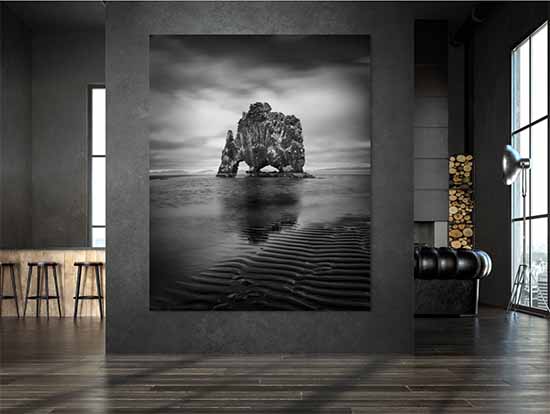 A large framed black-and-white vertical print of an Icelandic sea stack hanging on a dark concrete wall in a modern loft, beside a stacked firewood niche and tall windows.