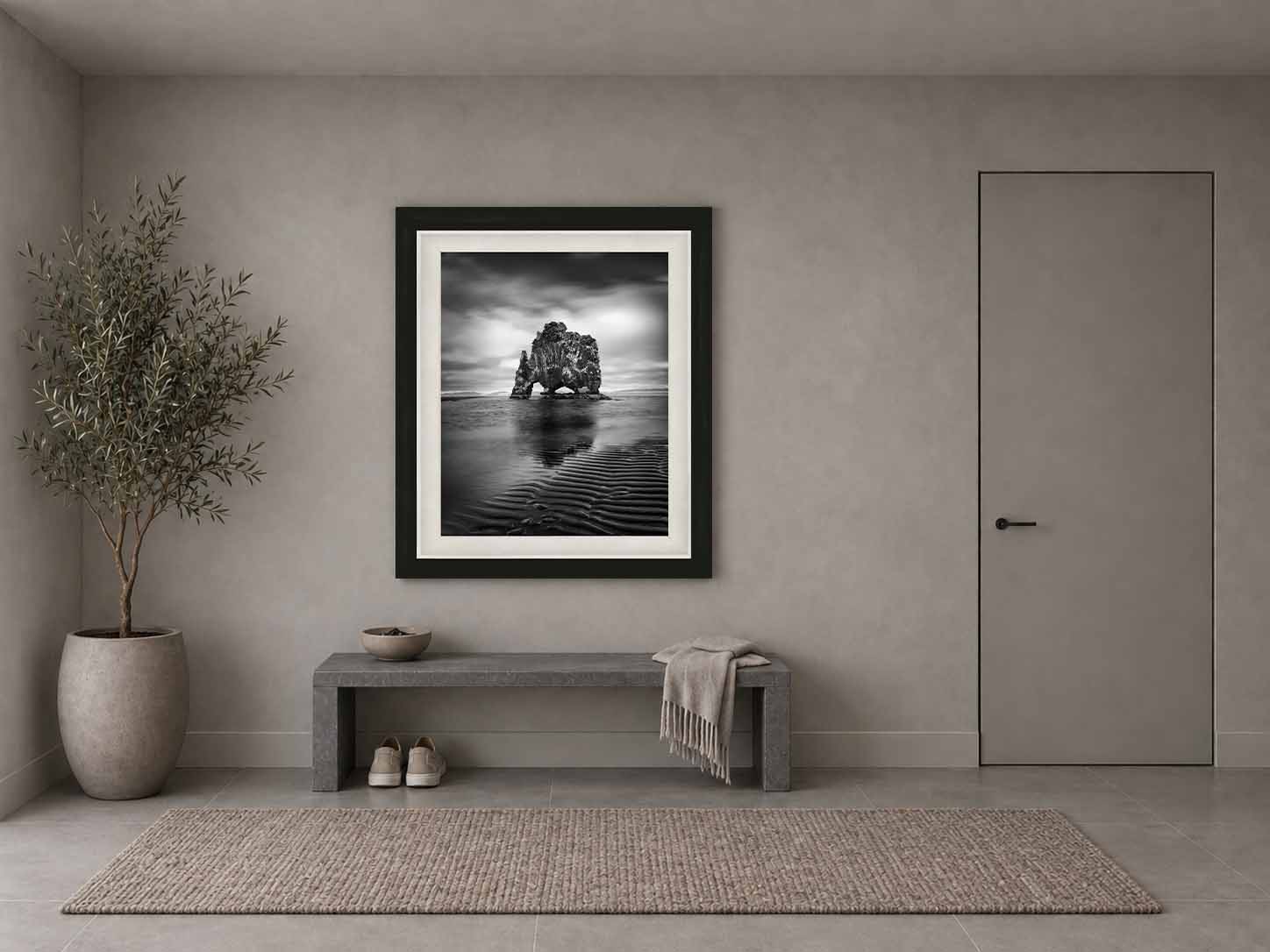 The Hv&iacute;tserkur sea stack at low tide in black and white, framed in black with a wide white liner above a stone bench in a neutral entry hall