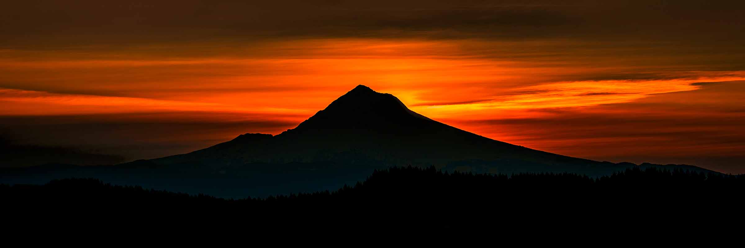 Hood in Shadow ARTIST’S STORY Mount Hood does not make promises. I spent an entire summer with a clear view of it from a friend's house, watching it every morning and every evening, learning its moods and its habits. Most days it was shrouded in cloud, its summit hidden behind grey that arrived before dawn and stayed well past dark. On the clearer days it rose against a featureless blue sky, beautiful in the way that familiar things are beautiful, present and real but not fully revealing itself. A mountain holding something back. That is the complicated relationship Hood has with those who love it. You can see it every day for weeks and still feel as if you have not quite seen it. As if the mountain is deciding, on its own terms and its own schedule, what it is willing to show you and when. And then one evening, without announcement or particular warning, the sky behind it caught fire. I watched it happen from the same place I had been watching every day all summer. The sun descended behind the horizon and the sky responded with a depth and intensity of color that I had not seen all season, deep crimson and burning orange spreading from horizon to horizon in horizontal bands that seemed to pulse with their own light. And against all of that, Mount Hood stood in complete and perfect darkness. Not diminished by the shadow but defined by it. Its geometry suddenly precise and absolute, the perfect triangle of its summit cutting into that burning sky with an authority that weeks of cloudy indifference had only sharpened. There is something profound about the relationship between shadow and light that this image understands completely. The mountain does not need to be illuminated to be present. Its darkness against that blazing sky is its own kind of statement, a reminder that sometimes the most powerful presence in a scene is the one that holds its form quietly while everything around it burns. I had watched that mountain every day for months. It waited until it was ready to show me this. Hood in Shadow is for anyone who has been patient with something that has not yet revealed its full beauty. Keep watching. The sky will eventually do what it did that evening. And when it does, you will understand that every ordinary day that came before it was simply the mountain deciding you were ready. Limited Fine Art Edition Each image is released in limited editions, with edition size set by presentation and scale, so the most substantial works remain the most rare.